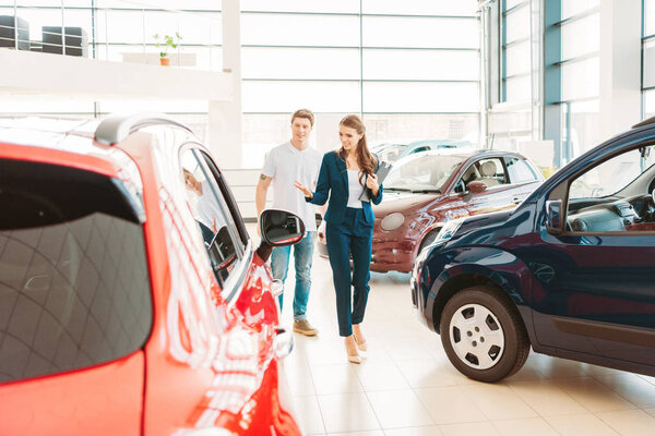 front view of woman with clipboard showing automobile to male client in car showroom 