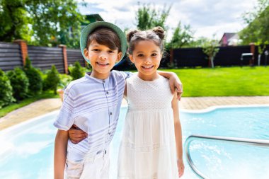 portrait of smiling little siblings hugging each other with swimming pool behind on summer day