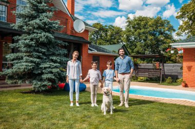 happy family with labrador dog looking at camera while holding hands on backyard of country house on summer day clipart
