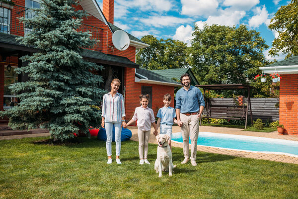 happy family with labrador dog looking at camera while holding hands on backyard of country house on summer day