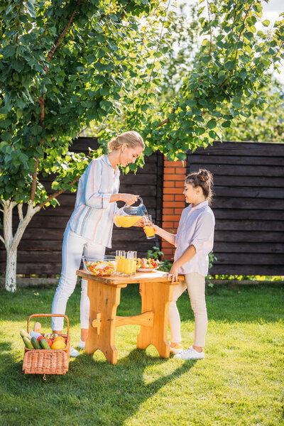 side view of smiling mother pouring juice into daughters glass during picnic on backyard