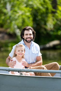 happy father and daughter sitting in boat on lake at park and looking at camera