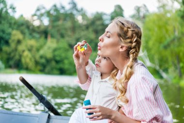 happy mother with son blowing soap bubbles while riding boat at park clipart