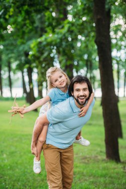 adorable daughter piggybacking on happy father and playing with toy plane at park clipart
