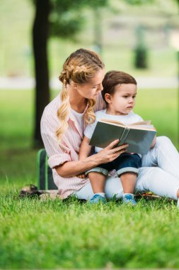 Anne ve oğlu Park'ta çimenlerin üzerinde kitapla oturan