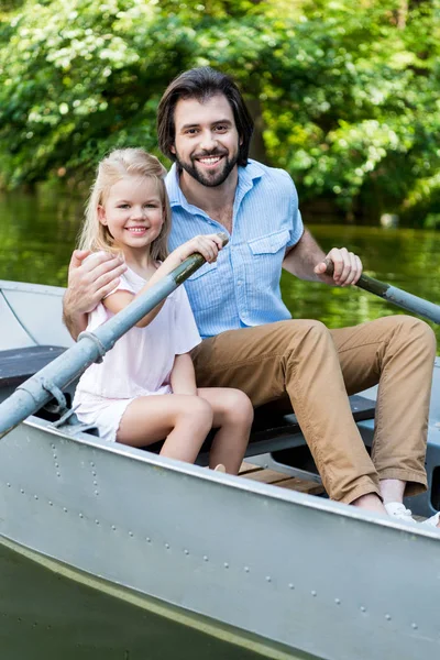 happy father and daughter riding boat and looking at camera on lake at park