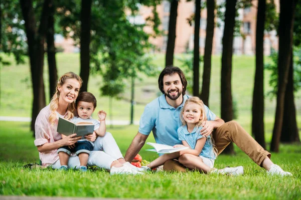 快乐的年轻家庭坐在草地上的书 看着相机 — 图库照片