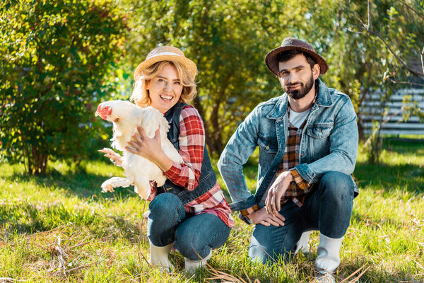 couple of farmers in straw hats sitting on grass with chicken outdoors 