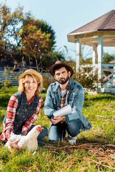 красивая улыбающаяся женщина держит курицу, в то время как ее парень делает заметки в бумагах на ферме
