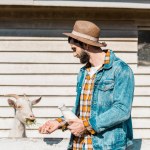 Side view of male farmer with bottle of milk feeding goat by grass near wooden fence at farm