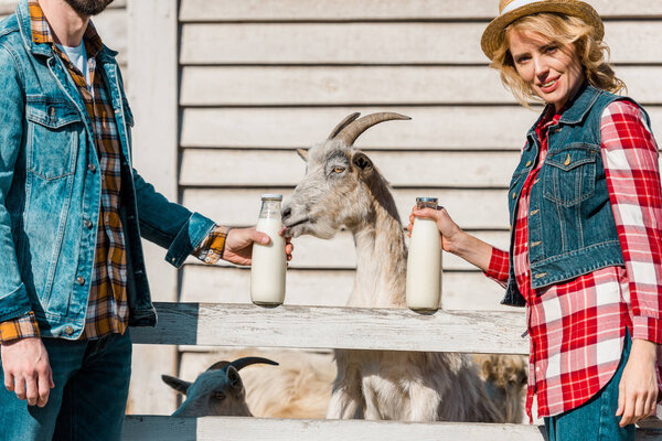 partial view of couple of farmers showing bottles of milk while goats grazing near wooden fence at ranch