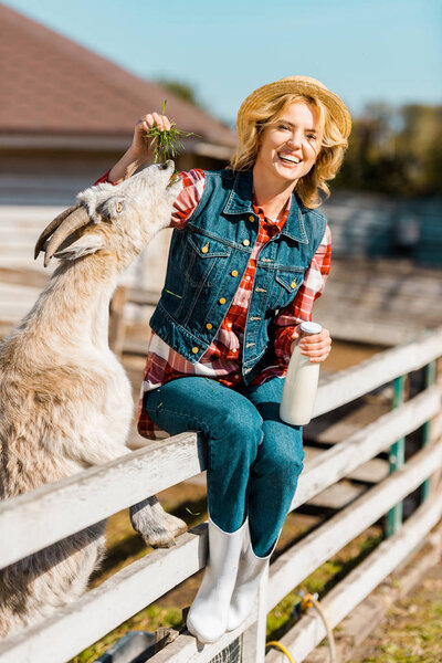 cheerful female farmer with milk bottle sitting on wooden fence and feeding goat at ranch