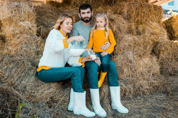 美丽的妇女倒入牛奶玻璃 而她的丈夫和女儿坐在农场附近 — 免费的图库照片