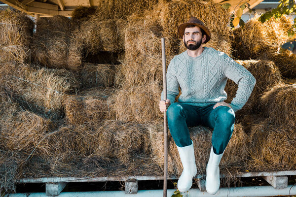 thoughtful male farmer in sweater and straw hat sitting on hay stacks at ranch