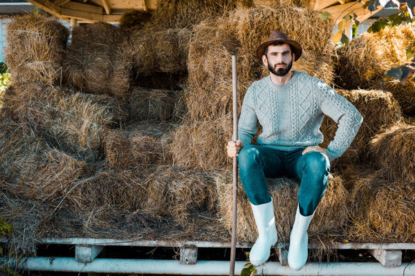 handsome male farmer in sweater and straw hat sitting on hay stacks at ranch