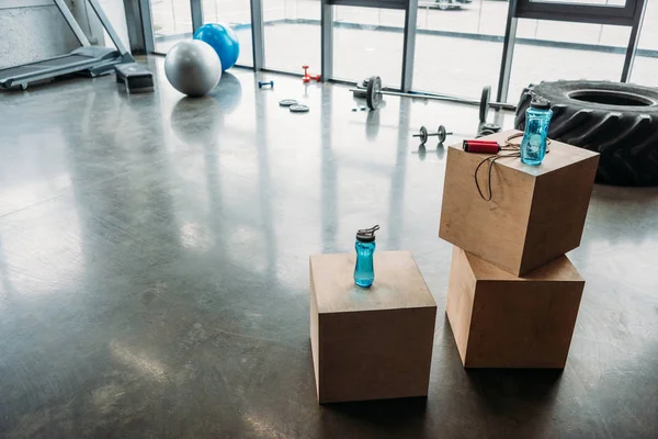 Cuerda de salto y botellas de agua en cajas de madera, mancuernas, barra de pesas, neumático de entrenamiento, bolas de fitness, plataforma de paso y cinta de correr en el gimnasio - foto de stock