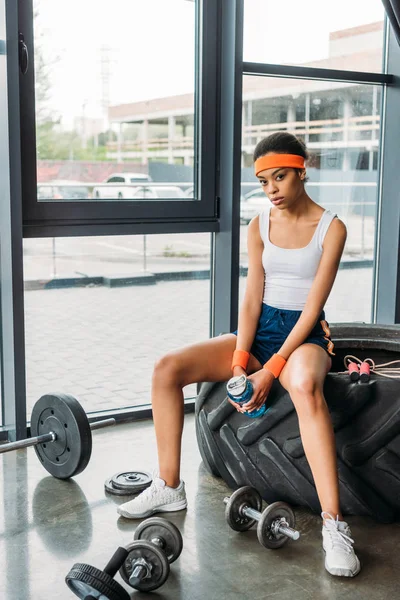 Jovem afro-americana esportista na cabeça e pulseiras sentado no pneu de treinamento com corda de salto e garrafa de água no ginásio — Fotografia de Stock