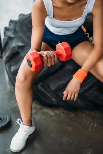 Abgeschnittenes Bild einer Sportlerin, die mit einer Hantel trainiert und im Fitnessstudio auf einem Trainingsreifen sitzt — Stockfoto