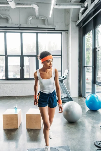 Jovem afro-americana esportista em headband e pulseiras exercitando-se na plataforma passo no ginásio — Fotografia de Stock