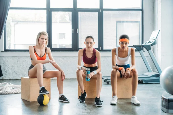 Trois sportives multiculturelles sérieuses assises sur des boîtes en bois au gymnase — Photo de stock