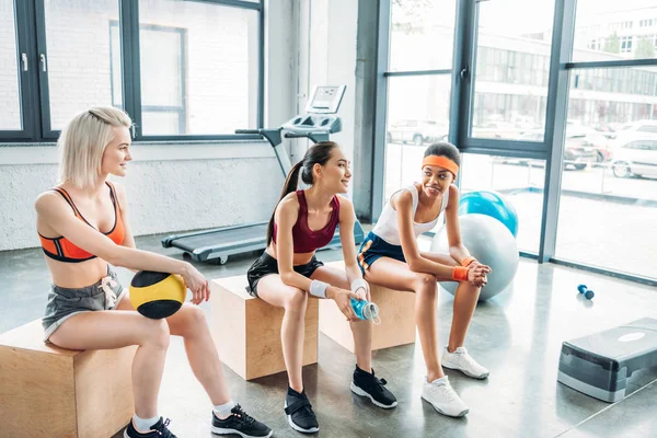 Young attractive multicultural sportswomen sitting on wooden boxes at gym — Stock Photo