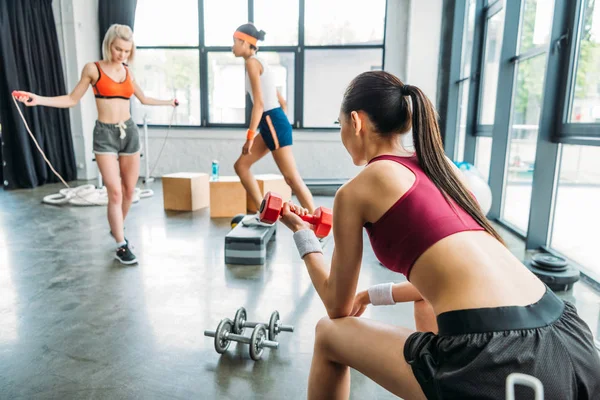 Jovem desportista fazendo exercício com haltere enquanto duas atletas do sexo feminino treinando atrás no ginásio — Fotografia de Stock