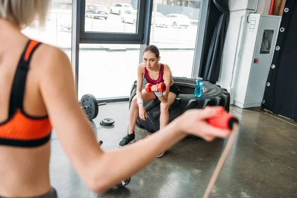 Recortado imagen de sportswoman haciendo salto cuerda entrenamiento mientras asiático femae atleta sentado en entrenamiento neumático con botella de agua y ejercicio con dumbbell en gimnasio - foto de stock