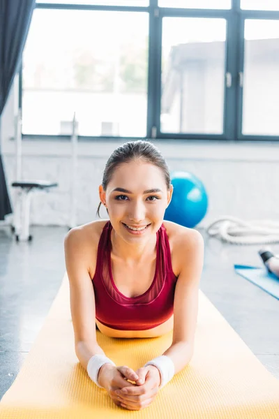 Sonriente asiático sportswoman en pulseras en fitness mat en gimnasio - foto de stock