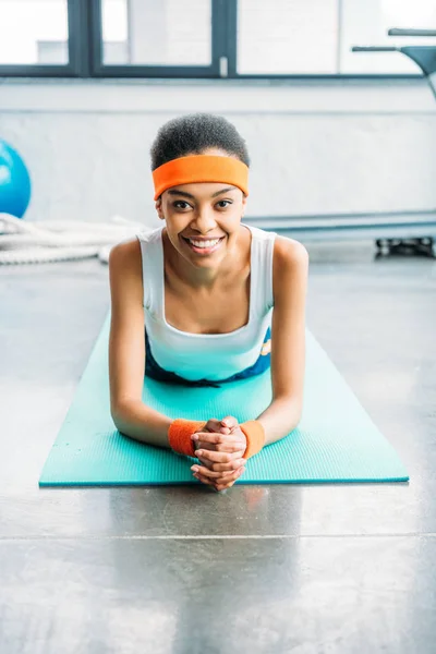 Smiling african american sportswoman in headband and wristbands laying on fitness mat at gym — Stock Photo