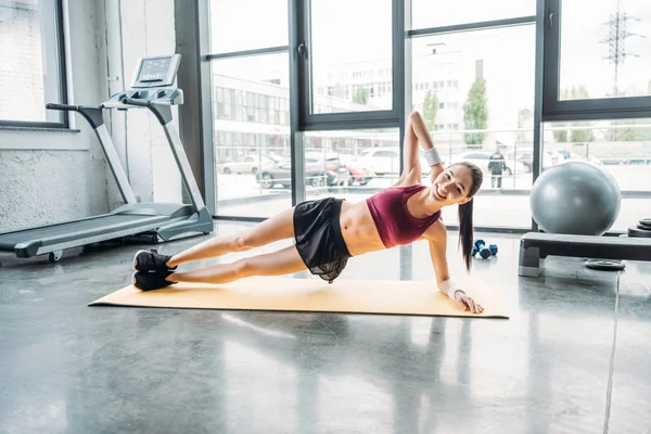 Sorridente asiatico sportswoman doing side plank su fitness mat a gym — Foto stock