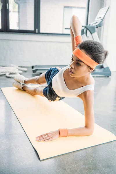 Young african american sportswoman in headband and wristbands doing side plank on fitness mat at gym — Stock Photo