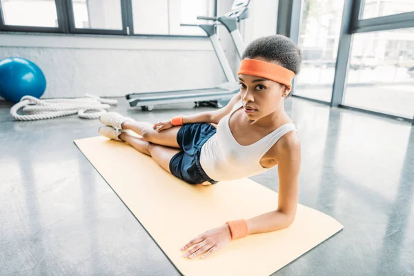 Atraente atleta afro-americano feminino exercitando-se no tapete de fitness e olhando para a câmera no ginásio — Fotografia de Stock