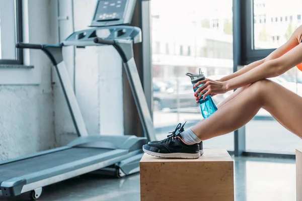 Cropped shot of sportswoman sitting on wooden box with bottle of water in front of treadmill at gym — Stock Photo