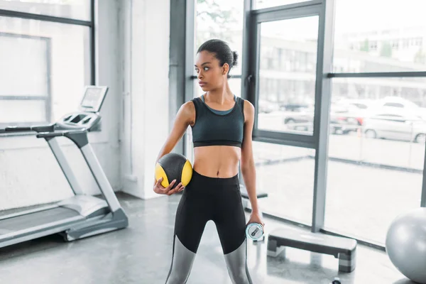 Ritratto di sportiva afro-americana con palla e borraccia sportiva in palestra — Foto stock