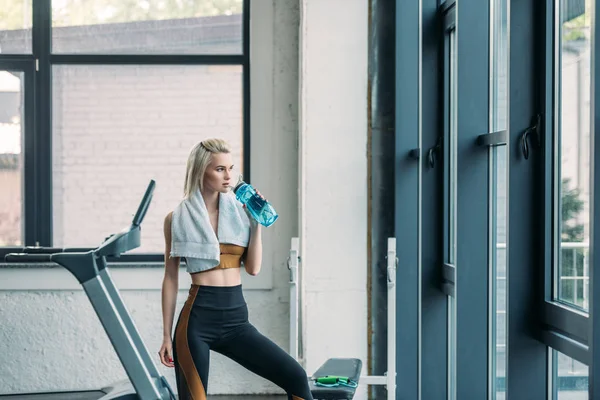 Young sportswoman with towel drinking water from sportive water bottle after workout at gym — Stock Photo