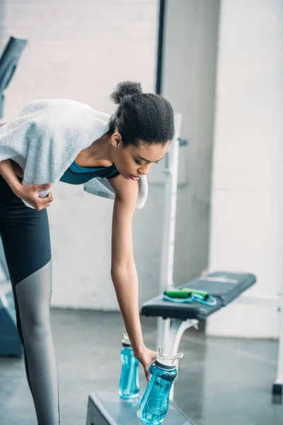 Deportista afroamericana con toalla que toma la botella de agua deportiva después del entrenamiento en el gimnasio - foto de stock