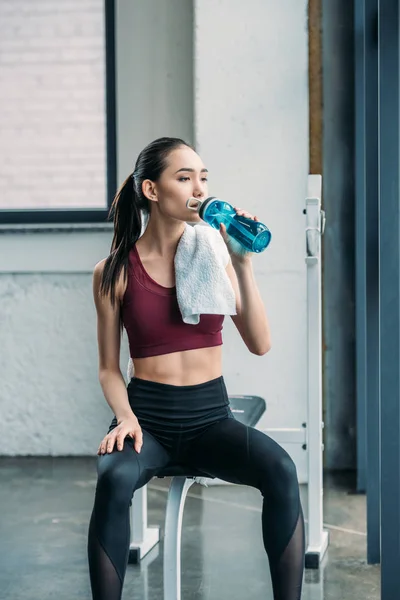 Asian sportswoman with towel drinking water from sportive water bottle after workout at gym — Stock Photo