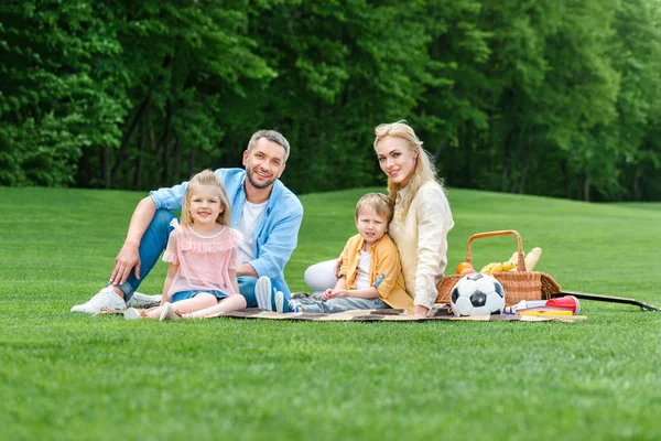 Familia feliz con dos niños sonriendo a la cámara mientras se sientan juntos en el picnic en el parque - foto de stock