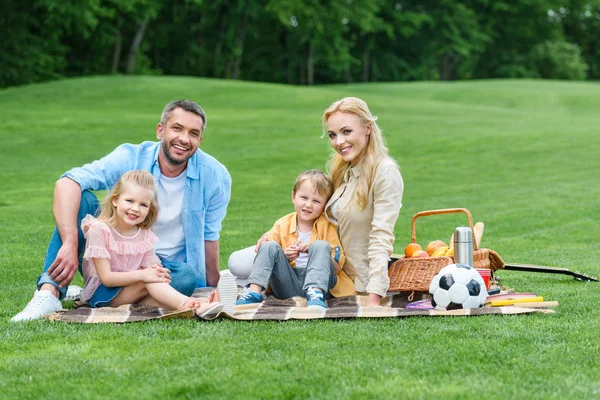 Famille heureuse avec deux enfants souriant à la caméra tout en étant assis ensemble sur plaid au pique-nique dans le parc — Photo de stock