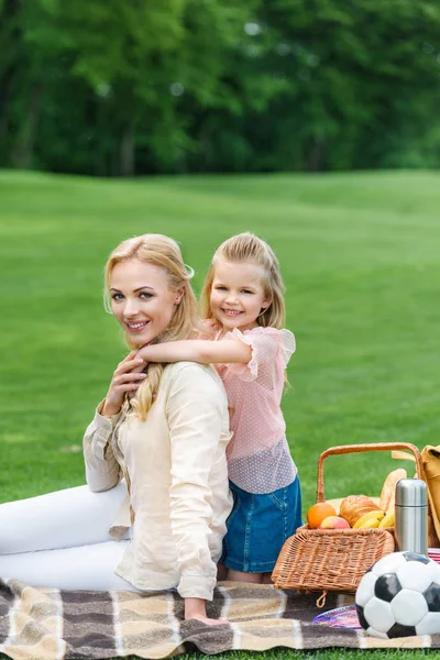 Feliz madre e hija abrazando y sonriendo a la cámara mientras se sientan juntos en el picnic a cuadros - foto de stock