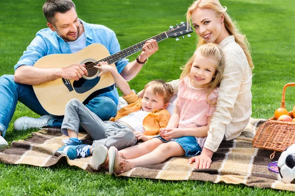 Familia feliz tocando la guitarra y sentados juntos a cuadros en el picnic - foto de stock