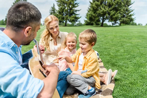 Happy family with guitar sitting together on plaid at picnic — Stock Photo
