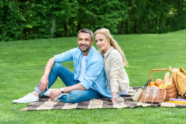 Happy couple sitting on plaid and smiling at camera in park — Stock Photo