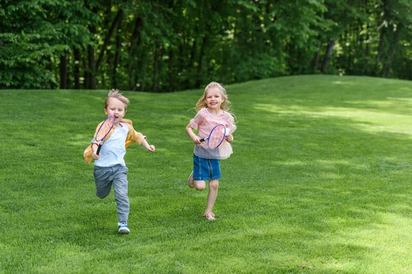 Adorables niños sonrientes con raquetas de bádminton corriendo juntos en el parque - foto de stock