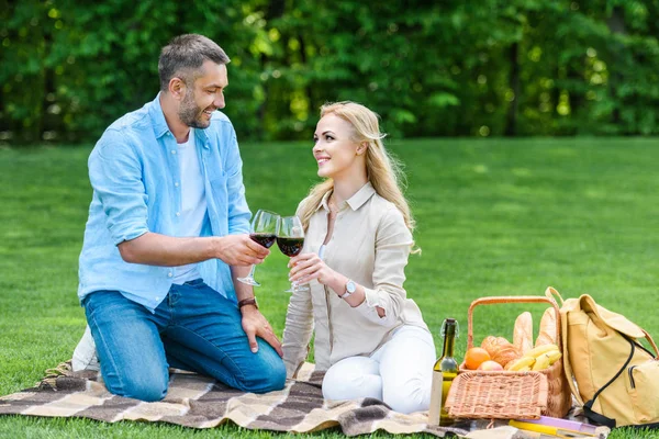 Happy couple clinking glasses of wine while sitting together on plaid at picnic — Stock Photo