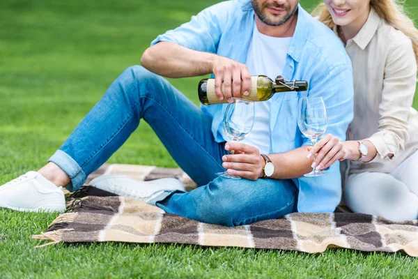 Cropped shot of couple pouring wine while sitting on plaid at picnic — Stock Photo