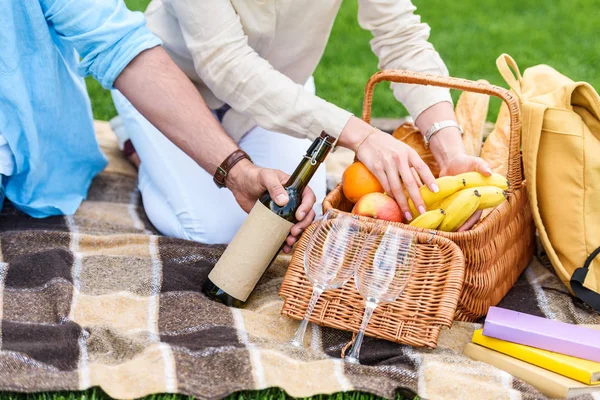 Cropped shot of couple sitting on plaid with wine and picnic basket — Stock Photo