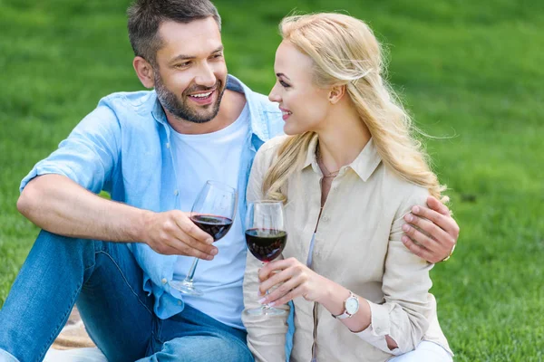 Happy young couple holding glasses of wine and smiling each other at picnic — Stock Photo