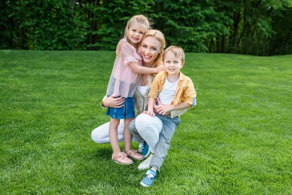 Glückliche Mutter umarmt entzückende Kinder und lächelt in die Kamera im Park — Stockfoto