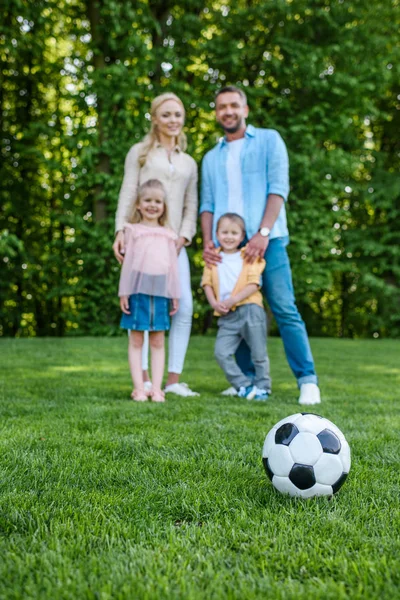 Vista de cerca de la pelota de fútbol en la hierba y la familia feliz de pie juntos en el parque - foto de stock